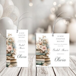 May include: Two white rectangular gift tags with floral arrangements and text. The tags read "A New Chapter Begins!" and "Bridal Shower" with the name "Olivia". The tags are tied with string and rest on a wooden surface.