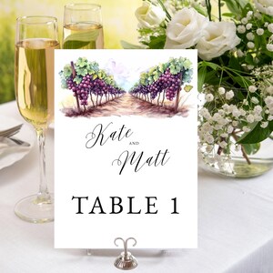 May include: A white table card with the names "Kate and Matt" and "TABLE 1" printed in black. The card features a watercolour illustration of a vineyard with purple grapes. A silver card holder supports the card.