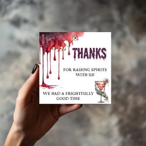 May include: Square card with a Halloween theme. The card features the word "THANKS" in purple, with red dripping blood-like accents. The text "FOR RAISING SPIRITS WITH US!" and "WE HAD A FRIGHTFULLY GOOD TIME" are also present.