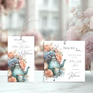 Può includere: Due etichette bianche per l'invito al brunch della festa della mamma con illustrazioni floreali e di teiere. Le etichette presentano il testo "Time for tea..." e "Mother's Day Brunch" con un nome. Le etichette sono legate con uno spago argentato.