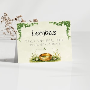 May include: A cream-colored card with the word "Lembas" in black script. The card features a gold ring surrounded by flowers and the text "Take one for the journey ahead."