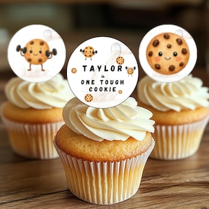 Può includere: Tre cupcake con glassa bianca e decorazioni commestibili. Le decorazioni presentano un biscotto dei cartoni animati con una faccia sorridente e il testo "TAYLOR is ONE TOUGH COOKIE". I cupcake sono in pirottini bianchi su una superficie di legno.