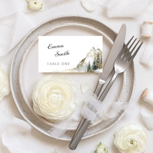 Op de afbeelding: Een elegante tafelsetting met een wit bord, bestek en een naamkaartje. De kaart toont "Emma Smith" en "TABLE ONE", met een aquarel berglandschap. Witte bloemen en linten maken de tafelsetting compleet.