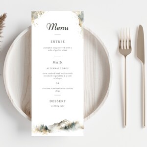 Peut inclure: Un menu de mariage avec une scène de montagne à l'aquarelle en bas. Le menu comprend des options d'entrée, de plat principal et de dessert. Une fourchette et un couteau dorés sont placés à droite d'une assiette blanche.