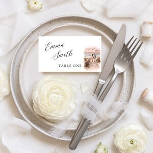 May include: A table setting featuring a white plate, cutlery, and a place card. The card displays "Emma Smith" and "Table One", with a watercolour illustration of a couple on a boardwalk. White flowers and ribbon add to the elegant aesthetic. The setting is ideal for a wedding.