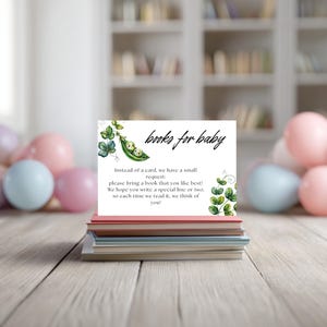 May include: A stack of books topped with a white card featuring the text "books for baby." The card has a green pea pod illustration with two baby faces inside. The card requests guests bring a book and write a special line.