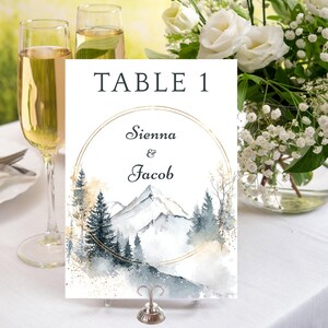 Könnte beinhalten: Eine Hochzeitstisch-Nummernkarte mit dem Text "TABLE 1". Die Karte zeigt ein Aquarelldesign von Bergen und Bäumen mit den Namen "Sienna & Jacob" in einem goldenen Kreis. Die Karte wird in einem silbernen Halter gehalten.