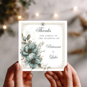 May include: A square wedding thank you card with a floral design and the text "Thanks for coming to the wedding of Brianna & Luke". The card is held by hands and features blue flowers and gold accents.