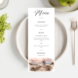 May include: A wedding menu card with the word "Menu" in elegant script. The menu lists entree, main course options, and dessert. A watercolour image of a couple walking on a beach is at the bottom. The card is on a white plate.