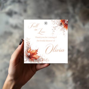 Può includere: Un biglietto d'invito quadrato con il testo "Fall in Love" e "Thank you for coming to the bridal shower of Olivia." Il biglietto presenta illustrazioni ad acquerello di foglie autunnali nei toni dell'arancione e del rosso.