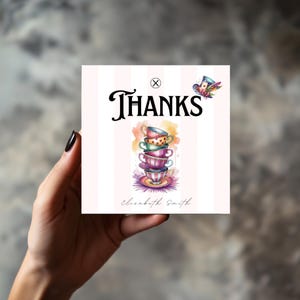 Puede incluir: Tarjeta cuadrada con una ilustración de acuarela de tazas de té apiladas en tonos rosa, morado, verde y naranja. La palabra "THANKS" está escrita en negro sobre las tazas. Una pequeña ilustración de sombrero está en la esquina superior derecha.