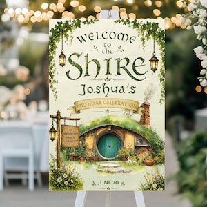 May include: A welcome sign for a birthday celebration. The sign reads "Welcome to the Shire, Joshua's Birthday Celebration" with an illustration of a hobbit hole. The sign is decorated with hanging lanterns and greenery.