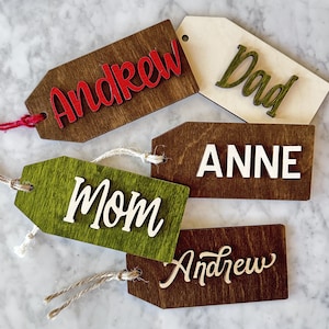 May include: Five wooden gift tags with names written on them. The tags are in various colors, including brown, green, and white. The names on the tags are Andrew, Dad, Anne, Mom, and Andrew.