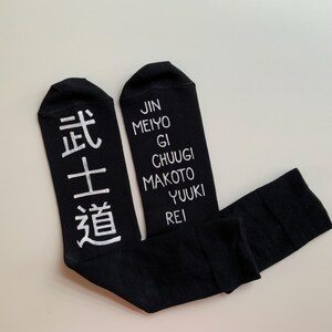 Pode incluir: Um par de meias pretas com texto branco. Uma meia tem os caracteres japoneses para "o caminho do guerreiro" e a outra meia tem as palavras "Jin Meiyo Gi Chuugi Makoto Yuuki Rei".