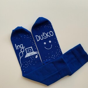 Op de afbeelding: Een paar blauwe sokken met witte letters. Eén sok zegt "Ing." met een cartoon-laptop en muis. De andere sok zegt "Duško" met een smiley.
