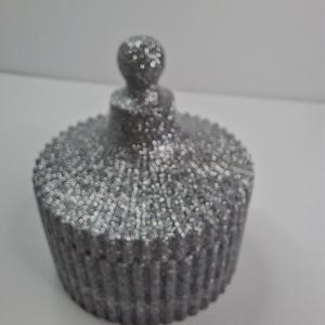 May include: A silver glitter-covered container with a lid. The container has a round, cupcake-like shape with a textured, ridged exterior. The lid is topped with a small, rounded knob. The overall effect is sparkly and decorative.