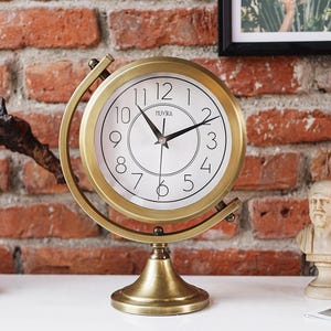 May include: A brass-colored globe clock with a white clock face and black numerals. The clock has a round face with the brand name "MUYIKA" printed at the top. The clock is set on a brass-colored stand.