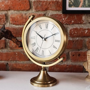 Puede incluir: Un reloj de globo de color latón con esfera blanca y números romanos negros. El reloj tiene una esfera redonda y está montado sobre un soporte de latón. El reloj está colocado contra una pared de ladrillo rojo.