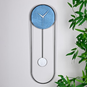 Könnte beinhalten: Eine moderne Wanduhr mit blauem, kreisförmigem Zifferblatt und weißen Zeigern. Die Uhr ist in einem schwarzen, länglichen Rahmen mit einem silberfarbenen Pendel eingefasst. Das Design ist minimalistisch und modern.