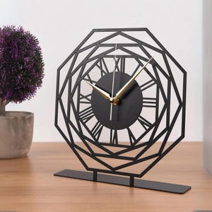 May include: Black tabletop clock with a geometric design and Roman numeral markings. The clock has gold-colored hands.