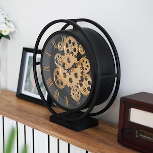 Può includere: Orologio nero e oro con ingranaggi e numeri romani. L'orologio presenta una cornice e una base nere, con un quadrante in vetro trasparente che rivela gli intricati ingranaggi dorati. Un oggetto decorativo.