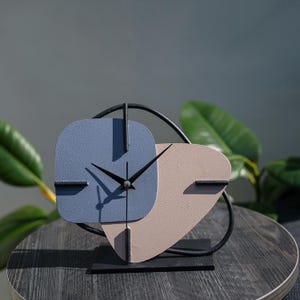 Puede incluir: Un reloj moderno y abstracto con una esfera azul y rosa y agujas negras. El reloj está sobre un soporte negro y tiene un marco de metal negro.
