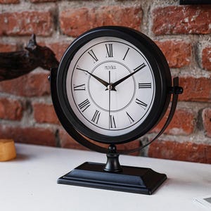 May include: A black analog clock with a white face and Roman numerals. The clock has a round shape and is supported by a black stand. The clock face has the brand name "MUYIKA" printed on it. The clock is a decorative item.