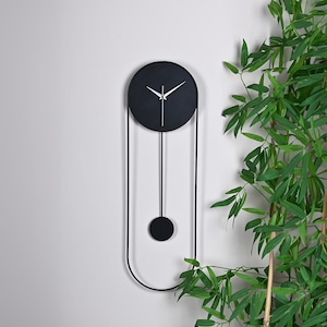 Könnte beinhalten: Eine moderne schwarze Wanduhr mit rundem Zifferblatt und weißen Zeigern. Die Uhr hat ein Pendeldesign mit einem schwarzen Ball am unteren Ende. Die Uhr ist an einer hellgrauen Wand montiert, mit grünen Pflanzen im Hintergrund.