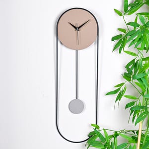 Op de afbeelding: Een moderne wandklok met een beige cirkelvormige wijzerplaat en zwarte wijzers. De klok hangt in een zwart metalen frame met een grijze slinger. Het ontwerp is minimalistisch en eigentijds, geschikt voor interieurdecoratie.