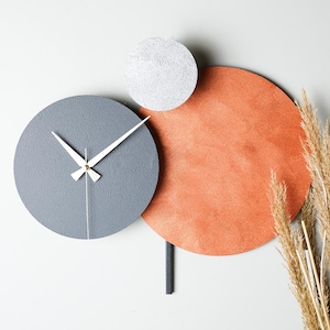 May include: A modern wall clock with three circular elements. The clock face is gray with white hands, the top element is silver, and the bottom element is orange.