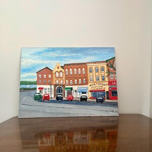 May include: An acrylic painting of a street scene with various buildings and cars. The buildings are in shades of brown, yellow, and red, with awnings and storefronts. The sky is blue with clouds. The painting is on a square canvas.