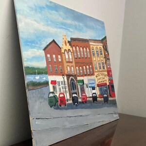 May include: An oil painting of a street scene with colorful buildings and small cars parked along the road. The sky is blue with white clouds. The painting includes the text "Northport Sweet Shop Luncheonette".