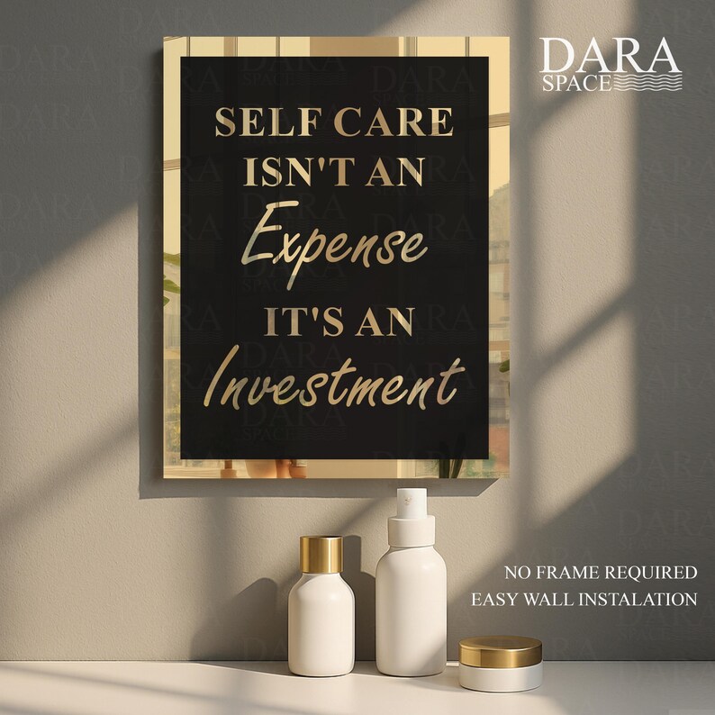 May include: A framed wall art print with the text "SELF CARE ISN'T AN Expense IT'S AN Investment" in gold lettering on a black background. The frame is gold. There are three white cosmetic bottles with gold lids in front of the art.