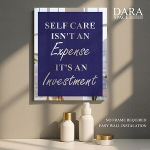 May include: A framed wall art print with the text "SELF CARE ISN'T AN Expense IT'S AN Investment" in silver lettering on a dark blue background. The image also shows white cosmetic bottles with gold lids.