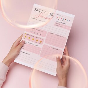 Pu&ograve; includere: Un diario di cura di s&eacute; rosa e bianco con le parole "SELF-CARE Journal" in elegante scrittura. Il diario include sezioni per la gratitudine, l'assunzione di acqua, il monitoraggio dell'umore, i pasti e le affermazioni. Il diario &egrave; tenuto da una persona.