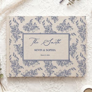 Puede incluir: Un álbum de fotos rectangular con una cubierta de tela beige que presenta un patrón floral azul. Un marco rectangular central contiene el texto The Smith, KEVIN & SOPHIA y March 9, 2026, impreso en tinta azul en la portada del libro.