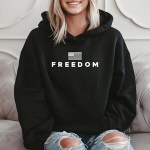 May include: Black hoodie with the word "FREEDOM" in white block letters. Above the word is a white graphic of the American flag. The hoodie has a drawstring hood and long sleeves.