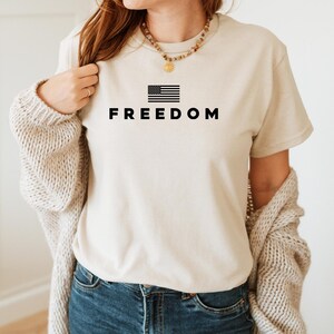 May include: A beige t-shirt with the word "FREEDOM" printed in black below a black and white American flag graphic. The shirt is worn with blue jeans and a beige cardigan.