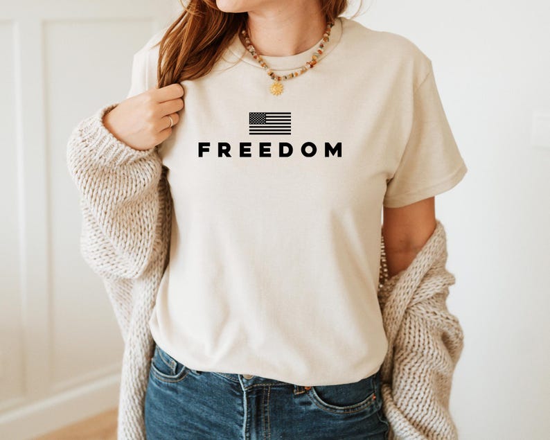 May include: A beige t-shirt with the word "FREEDOM" printed in black below a black and white American flag graphic. The shirt is worn with blue jeans and a beige cardigan.