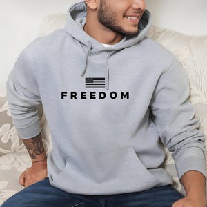 May include: A light gray hoodie with a front pocket and a hood. The word "FREEDOM" is printed in black block letters below a small black and white American flag graphic.