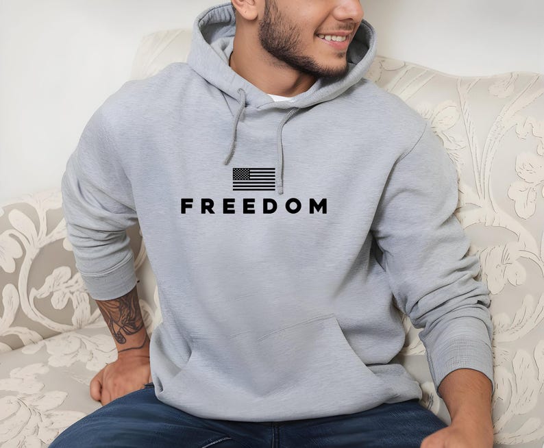 May include: A light gray hoodie with a front pocket and a hood. The word "FREEDOM" is printed in black block letters below a small black and white American flag graphic.