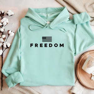 May include: A light teal hoodie with the word "FREEDOM" in bold black letters and a black and white American flag graphic. The hoodie has a drawstring hood and long sleeves.