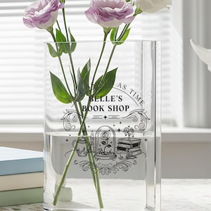 May include: A clear rectangular glass vase with water and flowers, featuring purple and white blooms. The vase has a black graphic that reads "Belle's Book Shop". A white angel statue and stacked books are also visible.