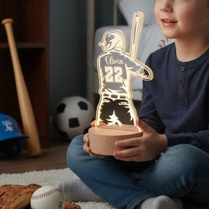 May include: A baseball-themed night light with the name "Olivia" and the number "22" illuminated. The light is a silhouette of a baseball player holding a bat, set on a wooden base. A baseball, glove, and bat are in the background.