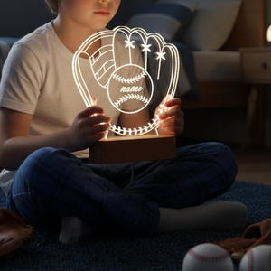 May include: A baseball-themed night light with a glove and ball design. The light is white and has the word "name" on the ball. The light sits on a wooden base. The overall theme is sports and home decor.