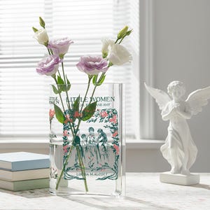 Puede incluir: Un jarrón rectangular de vidrio transparente con el texto "Little Women" y una ilustración floral. El jarrón contiene flores moradas y blancas. Una estatua de ángel blanco está a la derecha y libros a la izquierda.