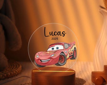 May include: A personalized night light with a red cartoon race car design. The clear circular panel displays the name "Lucas" and the year "2025". The light emanates from a wooden base, creating a warm glow.