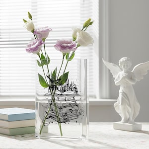 May include: A clear glass vase with a black graphic of books, a castle, and an owl, holding purple and white flowers. A white angel statue is on the right. Three books are stacked on the left.