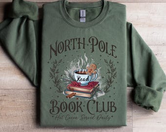 May include: An olive green sweatshirt with the words "North Pole Read Book Club" in a decorative font. The graphic includes a steaming mug, gingerbread man, books, and cinnamon sticks. The phrase "Hot Cocoa Served Daily" is also printed on the sweatshirt.