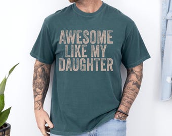 May include: Teal t-shirt featuring the text "AWESOME LIKE MY DAUGHTER" in a distressed, tan font. The shirt has short sleeves and a crew neck. The person is wearing blue jeans.